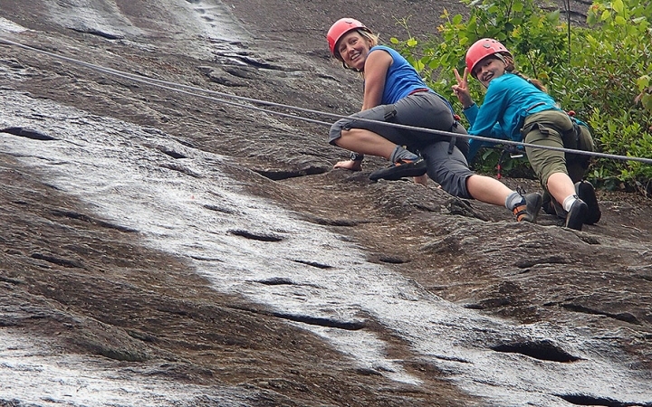 A parent and child look down and smile at the camera while climbing a rock wall. They are both wearing safety gear and are secured by ropes.